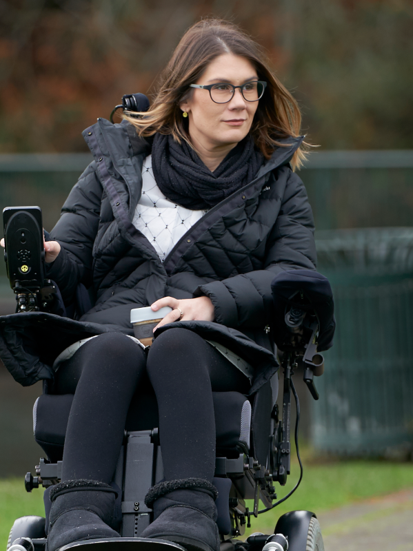 Women sits in her wheelchair holding a coffee outdoors.